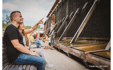 Ein junges Pärchen sitzt auf einer Bank am Gradierwerk im Kurpark Bad Sassendorf. Sie zeigt mit dem Finger in Richtung Gradierwerk und erläutert, warum die Sole am Gradierwerk herunterläuft.