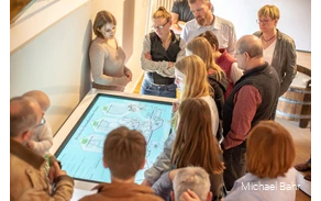 Eine Gruppe steht im Museum vor einem Bildschirm, ein Museumsguide erklärt etwas.