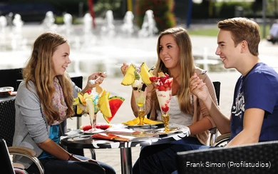 Drei Teenager sitzen im Eiscafé. Sie unterhalten sich und lachen. Vor ihnen auf dem Tisch stehen bunt dekorierte Eisbecher.